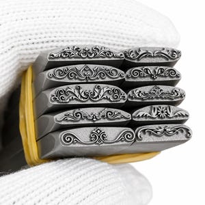 Op de afbeelding: Een set van tien metalen stempels met decoratieve bloemen- en krulontwerpen. De stempels worden bij elkaar gehouden door een gele band. De stempels zijn grijs en de ontwerpen zijn in het metaal ge&euml;tst.