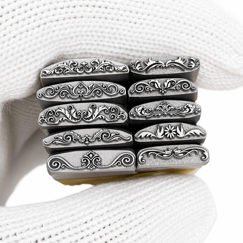 Op de afbeelding: Een verzameling metalen stempels met diverse decoratieve randontwerpen. Elke stempel heeft een ander sierlijk patroon, waaronder bloemen- en krulmotieven. De stempels worden vastgehouden in een witte gehandschoende hand, die de ingewikkelde details laat zien.