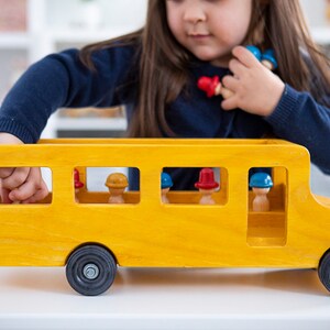 May include: A yellow wooden toy school bus with a black roof and wheels. The bus has windows with small wooden figures inside wearing red and blue hats.