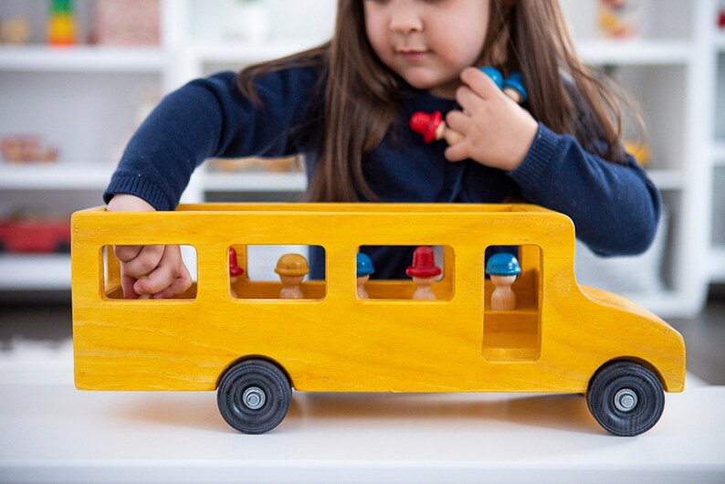 May include: A yellow wooden toy school bus with a black roof and wheels. The bus has windows with small wooden figures inside wearing red and blue hats.