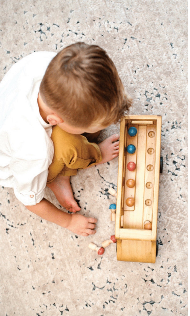 May include: A wooden toy truck with a removable top and a compartment for storing colorful balls. The truck has a blue, red, and brown color scheme. A child is playing with the truck and balls on a patterned rug.