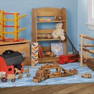 May include: A collection of wooden toys for children, including a red barn, a toy truck, a toy car carrier, a toy fire truck, a wooden toy train, a wooden toy helicopter, a wooden toy airplane, a wooden toy bear, a wooden toy bookshelf, a wooden toy marble run, and a wooden toy ramp. The toys are all made of natural wood and are painted in bright colors. The toys are arranged on a blue and white rug in a room with a blue wall and a window. The text "SAVING SHEPHERD" is visible on the wall.