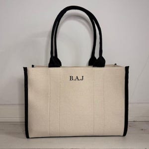 May include: Cream-colored tote bag with black trim and handles. The bag features the initials "B.A.J" in black letters on the front. The handles are thick and curved, and the bag has a rectangular shape.