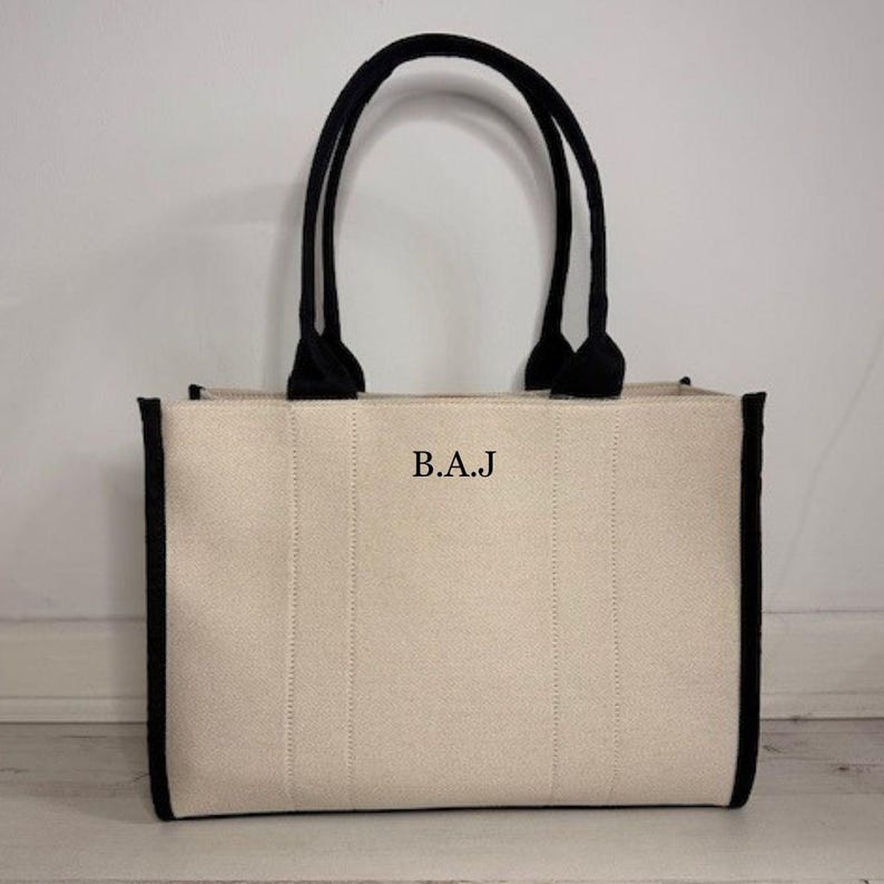 May include: Cream-colored tote bag with black trim and handles. The bag features the initials "B.A.J" in black letters on the front. The handles are thick and curved, and the bag has a rectangular shape.