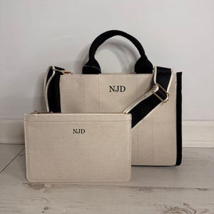 May include: A beige canvas tote bag and matching zippered pouch. The tote has black handles and a black and white striped shoulder strap. Both items have the initials "NJD" printed on them. The bag is rectangular and has a structured design.