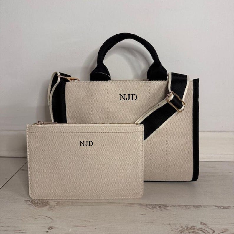 May include: A beige canvas tote bag and matching zippered pouch. The tote has black handles and a black and white striped shoulder strap. Both items have the initials "NJD" printed on them. The bag is rectangular and has a structured design.