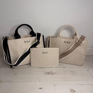 May include: Three beige canvas tote bags with black and beige straps. The bags have black and beige handles and the initials "B.A.J" and "H.M.F" printed on them. A matching pouch is also included.