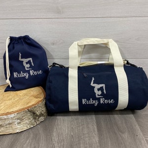 May include: A navy blue gym bag with off-white straps and a zip. The bag features a printed silver glitter design of a gymnast and a personalised name underneath. A smaller navy blue drawstring bag with the same design is also shown which can be purchased at an additional cost.