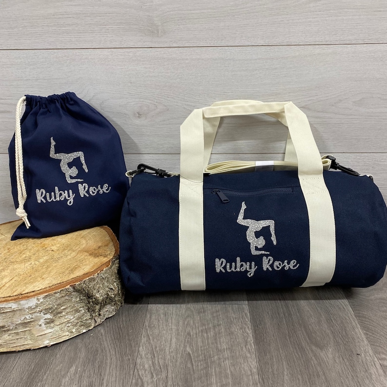 May include: A navy blue gym bag with off-white straps and a zip. The bag features a printed silver glitter design of a gymnast and a personalised name underneath. A smaller navy blue drawstring bag with the same design is also shown which can be purchased at an additional cost.