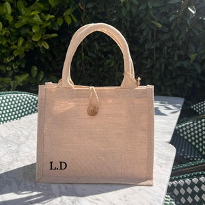 May include: A small, beige tote bag made of natural fibres with a wooden button closure. The bag has a simple design and is personalised with the initials "L.D."