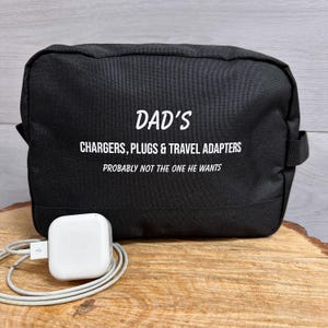 May include: Black travel bag with the text "DAD'S CHARGERS, PLUGS & TRAVEL ADAPTERS PROBABLY NOT THE ONE HE WANTS". A white charger with a grey cable is in front of the bag. The bag is on a wooden surface.