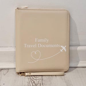 May include: A beige rectangular travel document organiser with a zip closure and a wrist strap. The front features the white text "Family Travel Documents" above a white line drawing of a heart and an aeroplane flying along a curved path.