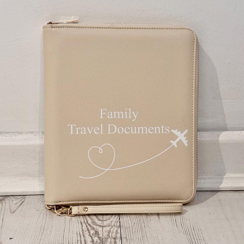 May include: A beige rectangular travel document organiser with a zip closure and a wrist strap. The front features the white text "Family Travel Documents" above a white line drawing of a heart and an aeroplane flying along a curved path.