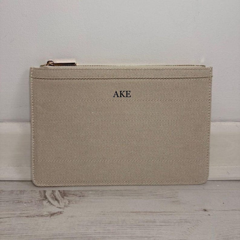 May include: A beige canvas zippered pouch with the initials "AKE" printed on the front. The pouch has a rectangular shape with a gold-colored zipper and a small, rectangular pull tab. The canvas material has a subtle diagonal weave.