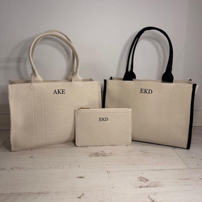 May include: Three canvas tote bags and a matching pouch. The bags are off-white with black handles and trim on one bag. The bags and pouch have embroidered initials. The initials "AKE" and "EKD" are visible on the bags and pouch.