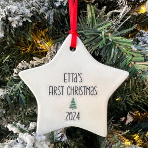 Peut inclure: Ornement de Noël en forme d'étoile en céramique blanche avec du texte noir qui dit "Etta's First Christmas 2024" et un petit graphique d'arbre de Noël vert.