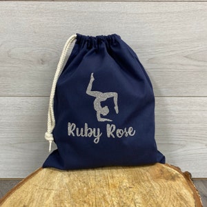 May include: A navy blue drawstring bag with a printed silver glitter design of a gymnast doing an elbow stand with a name personalised underneath in the same silver glitter.