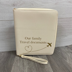 May include: A cream-coloured, rectangular travel document organiser with a gold zip closure and a matching wrist strap. The front features the gold-coloured text "Our family Travel documents" above a line drawing of a heart and an aeroplane.
