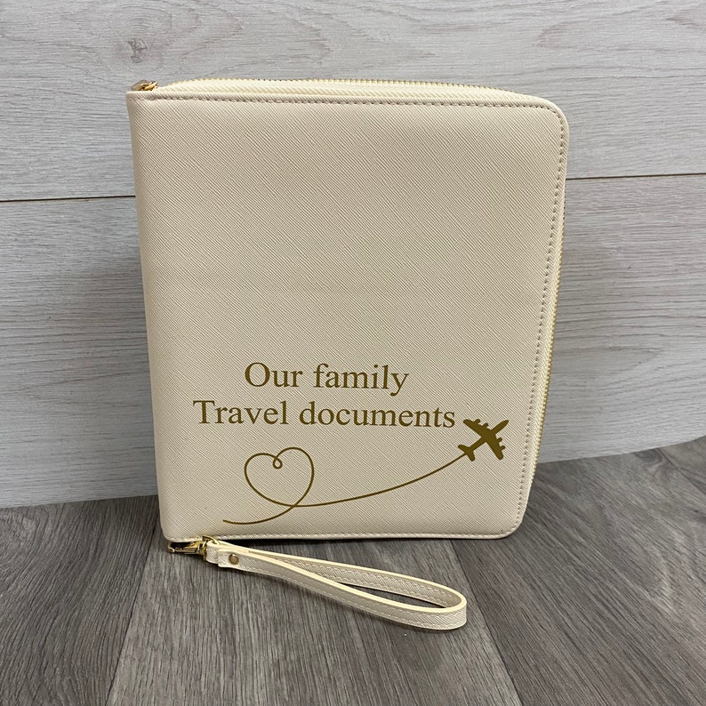 May include: A cream-coloured, rectangular travel document organiser with a gold zip closure and a matching wrist strap. The front features the gold-coloured text "Our family Travel documents" above a line drawing of a heart and an aeroplane.