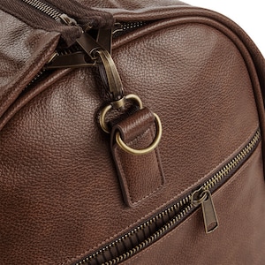 Pu&ograve; includere: Primo piano di una borsa da viaggio in pelle marrone con una tasca con cerniera e un tirare della cerniera in ottone. La borsa ha una finitura in pelle testurizzata.