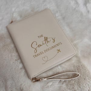 May include: A cream-coloured travel document holder with a zip closure and wrist strap. The front displays the text "The Smith's Travel Documents" in gold script, accompanied by an airplane and heart design.