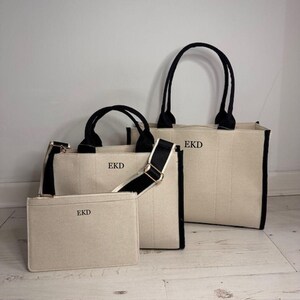 May include: A set of three canvas bags in varying sizes. The bags are off-white with black trim and handles. Each bag has the initials "EKD" printed on the front. One bag has a shoulder strap.