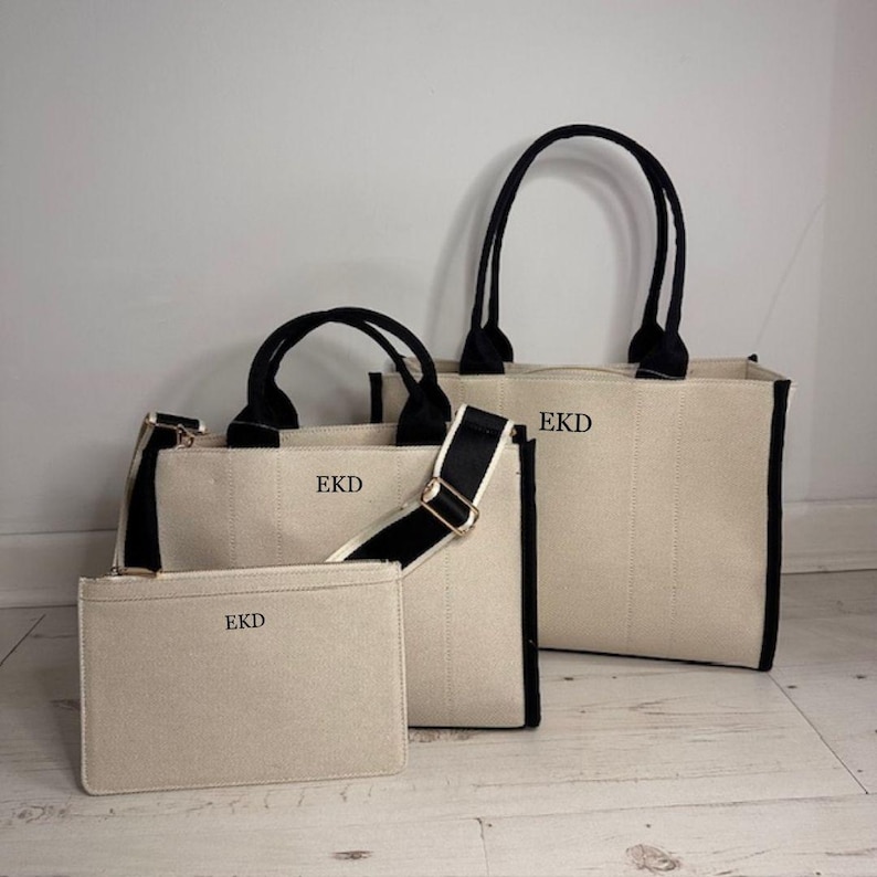 May include: A set of three canvas bags in varying sizes. The bags are off-white with black trim and handles. Each bag has the initials "EKD" printed on the front. One bag has a shoulder strap.