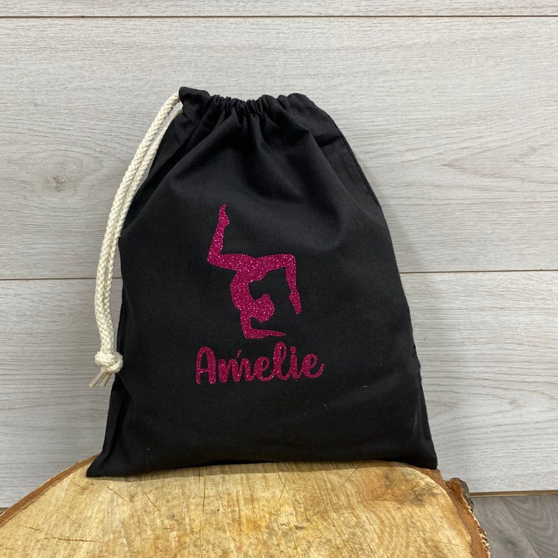 May include: Black drawstring bag with a pink glitter gymnast silhouette and a personalised name underneath the gymnast in pink glitter.