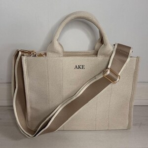 May include: Beige canvas tote bag with a matching adjustable shoulder strap and top handles. The bag features the letters "AKE" printed on the front. The bag has a gold-colored zipper and hardware.