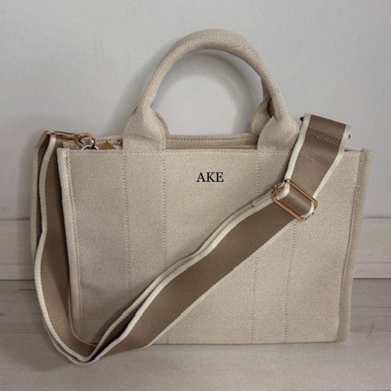 May include: Beige canvas tote bag with a matching adjustable shoulder strap and top handles. The bag features the letters "AKE" printed on the front. The bag has a gold-colored zipper and hardware.