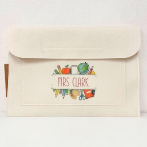 May include: A cream-coloured felt envelope-style case with a flap closure. The front displays a colourful illustration of school supplies, including pencils, an apple, a globe, and the name "MRS CLARK" in pink.