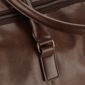 Pu&ograve; includere: Primo piano di una borsa da viaggio in pelle marrone con una fibbia in metallo dorato. La borsa ha una chiusura a zip e una finitura in pelle testurizzata.