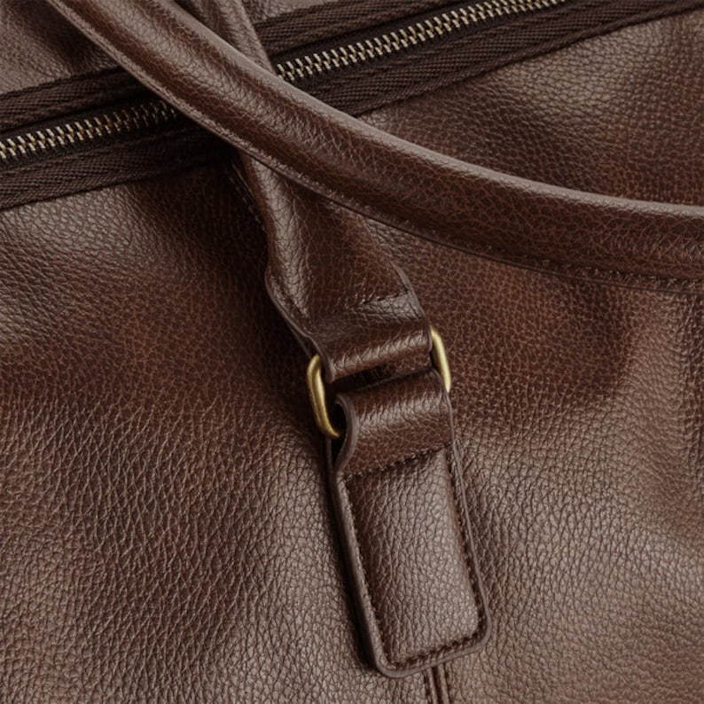 Pu&ograve; includere: Primo piano di una borsa da viaggio in pelle marrone con una fibbia in metallo dorato. La borsa ha una chiusura a zip e una finitura in pelle testurizzata.