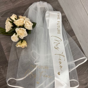 Peut inclure: Un ensemble de mariée comprenant un voile blanc, une écharpe en satin avec l'inscription dorée "THE FUTURE Mrs. Fenn" et un bouquet de roses crème. Le voile porte également du texte doré. Un cœur doré est inclus.