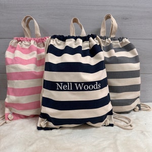 May include: Three striped drawstring bags in pink and white, navy and cream, and grey and cream. The navy bag features the text "Nell Woods" in white. Each bag has cream drawstrings and handles. The bags are made of fabric.