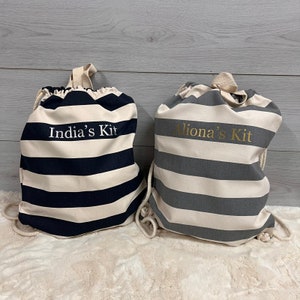 May include: Two drawstring bags with horizontal stripes. One bag is navy blue and cream, with "India's Kit" printed on it. The other bag is gray and cream, with "Aliona's Kit" printed on it. Both bags have cream-colored drawstrings and handles.