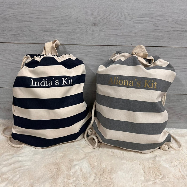 May include: Two drawstring bags with horizontal stripes. One bag is navy blue and cream, with "India's Kit" printed on it. The other bag is gray and cream, with "Aliona's Kit" printed on it. Both bags have cream-colored drawstrings and handles.