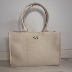 May include: A beige canvas tote bag with two handles. The bag features the initials "CJJ" in the center. Vertical stitching details the front of the bag.