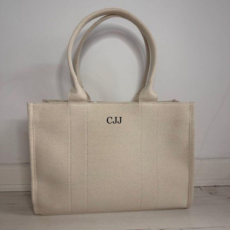 May include: A beige canvas tote bag with two handles. The bag features the initials "CJJ" in the center. Vertical stitching details the front of the bag.