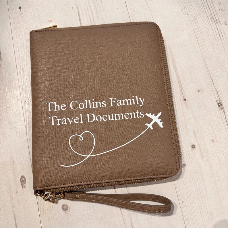 May include: A taupe-coloured zipped travel document organiser featuring a wrist strap. The front is printed with the white text "The Collins Family Travel Documents" accompanied by a graphic of an aeroplane flying along a heart-shaped path.