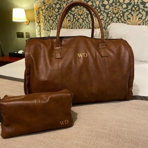 Pu&ograve; includere: Una borsa da viaggio in pelle marrone con un beauty case in pelle marrone abbinato. Entrambe le borse hanno le iniziali "WD" ricamate in filo dorato.