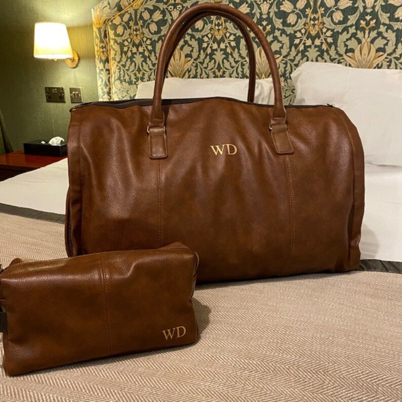 Pu&ograve; includere: Una borsa da viaggio in pelle marrone con un beauty case in pelle marrone abbinato. Entrambe le borse hanno le iniziali "WD" ricamate in filo dorato.