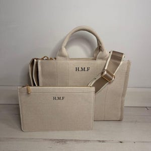 May include: Beige canvas tote bag with a matching zippered pouch. The tote features a top handle, a detachable shoulder strap, and the initials "H.M.F" printed on the front. The pouch also has the initials "H.M.F".