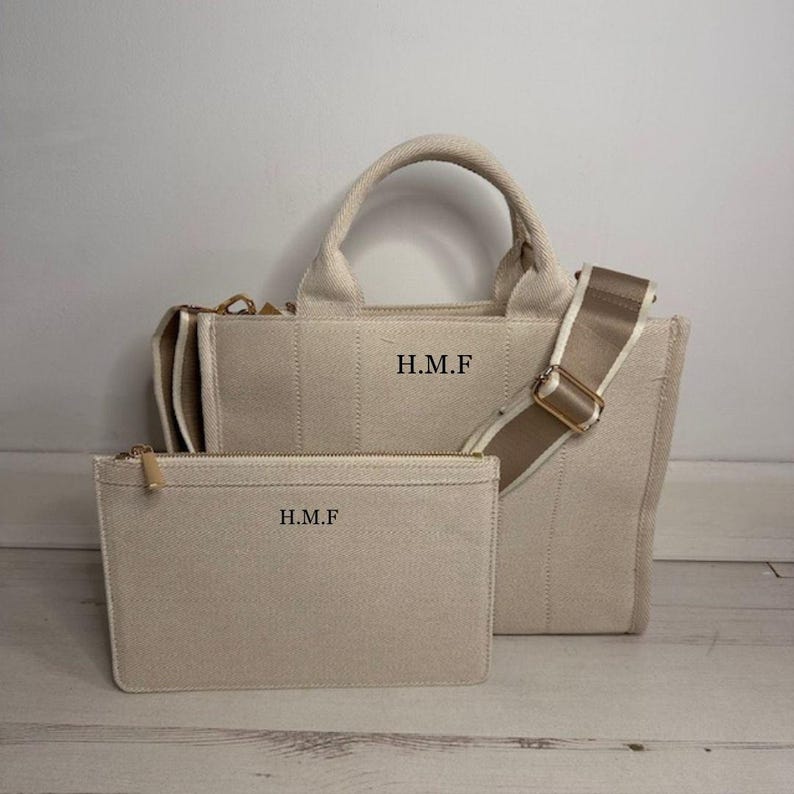 May include: Beige canvas tote bag with a matching zippered pouch. The tote features a top handle, a detachable shoulder strap, and the initials "H.M.F" printed on the front. The pouch also has the initials "H.M.F".