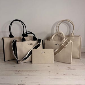 May include: A collection of beige canvas tote bags and a matching pouch. The bags have black or beige handles and straps, with the names "ELLE" and "AKE" printed on them. The bags are of different sizes.