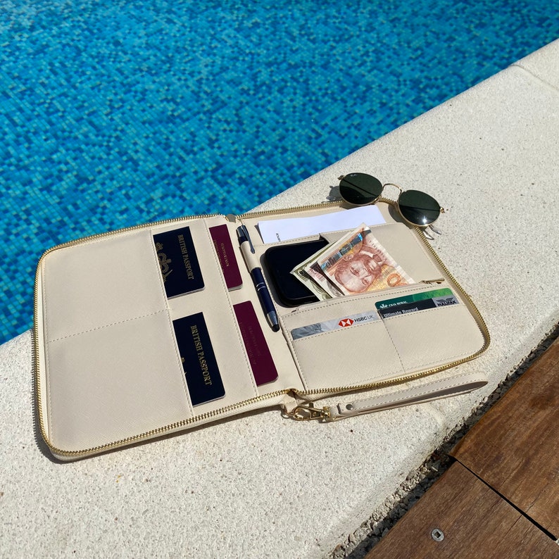 May include: A cream-coloured zippered travel organiser wallet featuring multiple interior slots containing two dark blue British passports, two burgundy passports, a black smartphone, a pen, paper currency, and several credit cards.