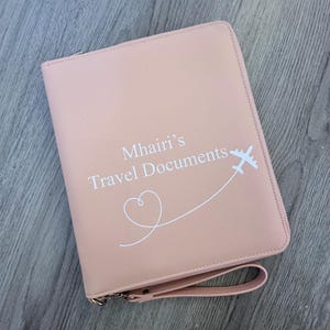 May include: A light pink rectangular travel document organiser with a zip closure and a wrist strap. The front features white text that reads Mhairi's Travel Documents, accompanied by a white graphic of an aeroplane and a heart-shaped flight path.