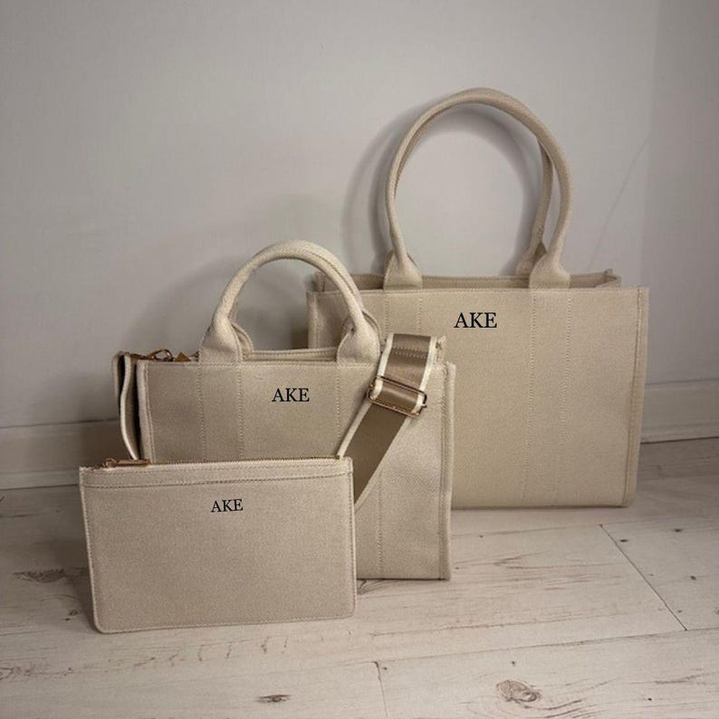 May include: Three beige canvas bags of varying sizes, including a tote bag, a shoulder bag with a strap, and a small zippered pouch. Each bag features the initials "AKE" printed on the front.