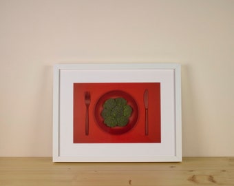 Pode incluir: Uma impressão emoldurada com fundo vermelho, um prato de brócolis verde, um garfo e uma faca. Os talheres são vermelhos como o fundo. A moldura é branca, sobre uma superfície de madeira. Arte culinária minimalista.