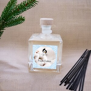 Könnte beinhalten: Eine klare Glasflasche mit einem Holzdeckel und einem Etikett mit der Aufschrift "Qiss Wax Fresh Cotton". Die Flasche ist mit einer hellfarbigen Flüssigkeit gefüllt und steht auf einer hellbraunen Oberfläche. Im Hintergrund befinden sich schwarze Diffusorstäbchen.